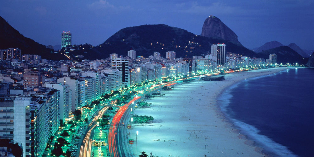 Copacabana beach at dusk with flickering building lights and sugarloaf mountain in the distance